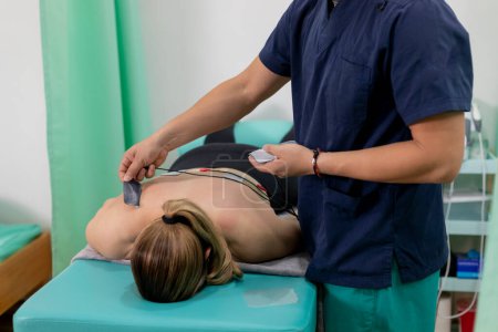 Foto de Fisioterapeuta colocando almohadillas de electrodos en la espalda de una mujer que está acostada en una camilla - Imagen libre de derechos