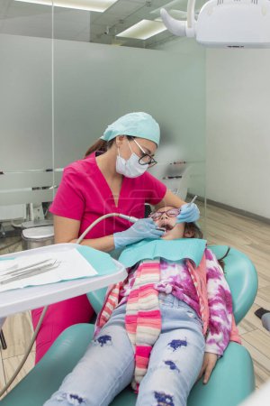 Photo for Vertical image of a dentist cleaning the teeth of a young girl lying on a chair in the dental office. - Royalty Free Image