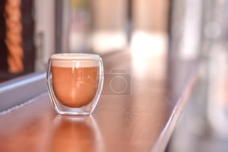 A transparent glass of latte stands outdoors on the tabletop of a coffee shop