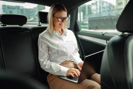Woman accountant working on laptop sitting car leather backseat on way to office. High quality photo