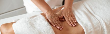 Slim young woman lying on massage table under white towels while masseuse massaging her abdomen