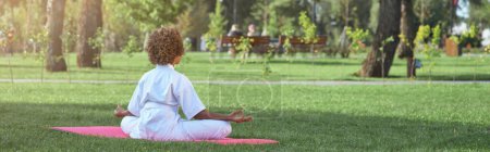Téléchargez les photos : Vue arrière de l'enfant afro-américaine assise sur un tapis de yoga en position lotus et faisant de l'exercice de méditation à l'extérieur - en image libre de droit