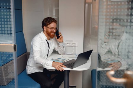 Foto de Jefe masculino hablando de teléfono con el cliente mientras está sentado en el espacio de coworking y trabajando en el ordenador portátil - Imagen libre de derechos