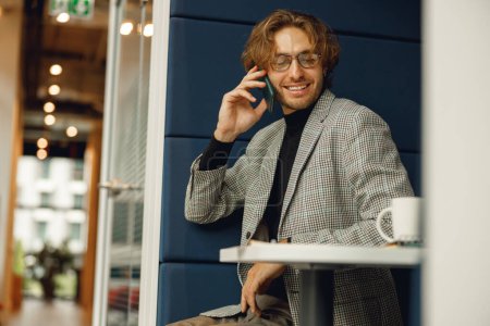 Foto de Jefe masculino hablando de teléfono con el cliente mientras está sentado en el espacio de coworking y trabajando en el ordenador portátil - Imagen libre de derechos