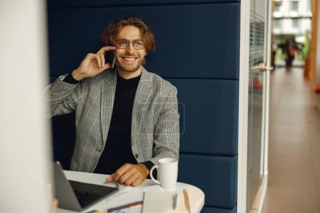 Foto de Empresario hablando de teléfono con el cliente mientras está sentado en coworking y trabajando en el ordenador portátil - Imagen libre de derechos