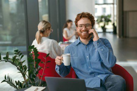 Foto de Freelancer masculino hablando por teléfono con el cliente mientras está sentado en coworking y trabajando en el portátil - Imagen libre de derechos