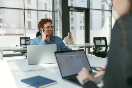 Foto de Empresario hablando teléfono con el cliente mientras está sentado en la oficina moderna y trabajando en el ordenador portátil - Imagen libre de derechos