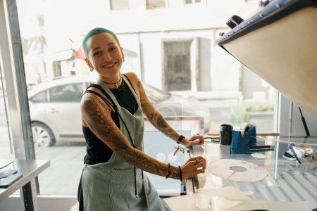 Trabajadora sonriente en delantal usando máquina de impresión en un taller. Foto de alta calidad