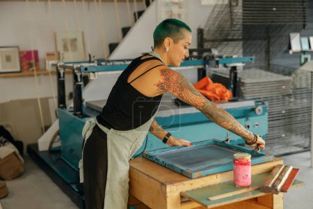 Female worker with squeegee is pressing ink on frame working in design studio. Silk screen printing