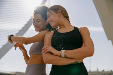 Téléchargez les photos : Femme souriante avec une amie regardant smartwatch avant de faire de l'exercice debout à l'extérieur - en image libre de droit