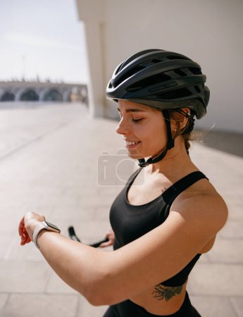 Téléchargez les photos : Femme souriante en tenue de sport regardant smartwatch après le vélo. Sports de plein air le matin - en image libre de droit