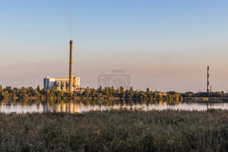 The old waste processing plant on the lake bank emits air pollution from its smoking smokestacks. It harms the environment and creates an unsightly backdrop against a colorful sunset sky.