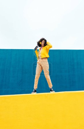 Foto de Joven mujer feliz bailando y divirtiéndose al aire libre. Adolescente escuchando música con smartphone y auriculares en una zona urbana moderna de color amarillo y azul - Imagen libre de derechos