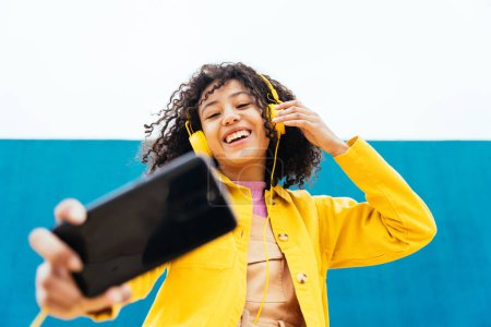 Foto de Joven feliz mujer tomando selfies y divertirse al aire libre. Adolescente escuchando música con smartphone y auriculares en una zona urbana moderna de color amarillo y azul - Imagen libre de derechos