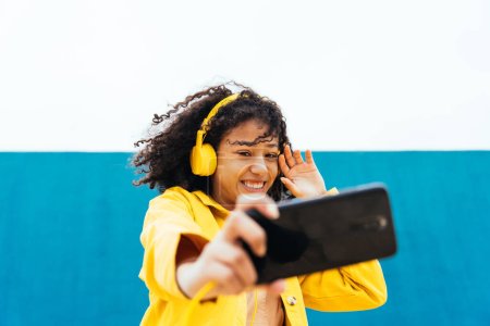 Téléchargez les photos : Jeune femme heureuse prenant selfies et s'amusant en plein air. Adolescent écoutant de la musique avec smartphone et écouteurs dans une zone urbaine moderne jaune et bleue - en image libre de droit