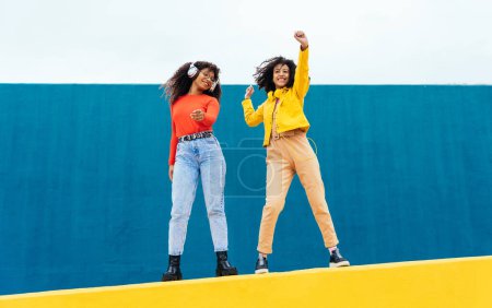 Foto de Mujeres jóvenes felices bailando y divirtiéndose al aire libre. Adolescentes escuchando música con smartphone y auriculares en una zona urbana moderna de color amarillo y azul - Imagen libre de derechos