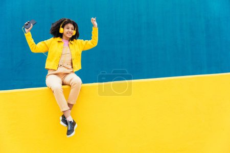 Téléchargez les photos : Jeune femme heureuse dansant et s'amusant en plein air. Adolescent écoutant de la musique avec smartphone et écouteurs dans une zone urbaine moderne jaune et bleue - en image libre de droit
