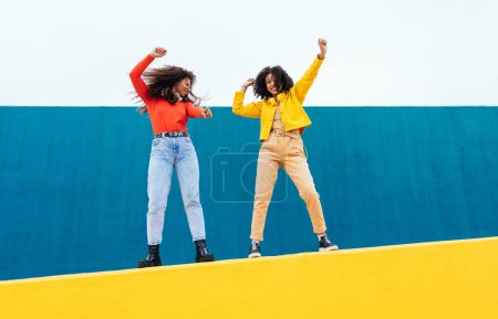 Photo for Young happy women dancing and having fun outdoor. Teenagers listening to music with smartphone and headphones in a yellow and blue modern urban area - Royalty Free Image