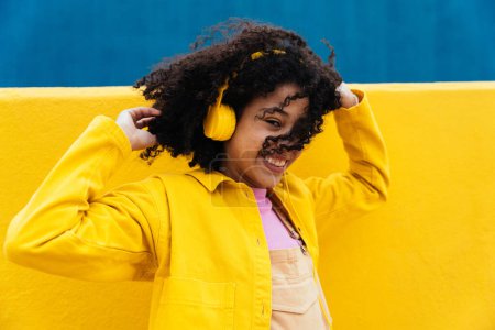 Photo for Young happy woman dancing and having fun outdoor. Teenager listening to music with smartphone and headphones in a yellow and blue modern urban area - Royalty Free Image