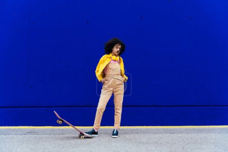 Téléchargez les photos : Jeune femme heureuse s'amuser en plein air avec son skateboard. Adolescent écoutant de la musique avec smartphone et écouteurs dans une zone urbaine moderne jaune et bleue - en image libre de droit
