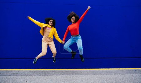 Téléchargez les photos : Jeunes femmes heureuses dansant et s'amusant en plein air. Les adolescents écoutent de la musique avec smartphone et écouteurs dans une zone urbaine moderne jaune et bleue - en image libre de droit