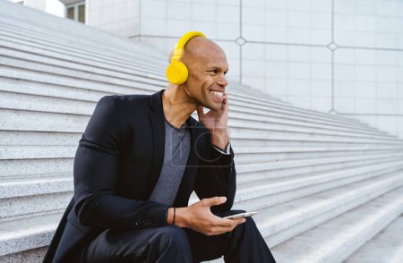 Photo for Young business man sitting and listening to music in the financial street area of the city .Representation of a successful sales person reaching the top of the career. - Royalty Free Image