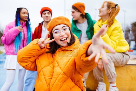 Foto de Grupo multirracial de jóvenes amigos felices que se reúnen al aire libre en invierno, vistiendo chaquetas de invierno y divirtiéndose, mujer asiática haciendo gestos en primer plano - Millennials multiétnicos que se unen en un área urbana, conceptos sobre la juventud y las liberaciones sociales - Imagen libre de derechos