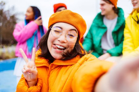 Foto de Grupo multirracial de jóvenes amigos felices que se reúnen al aire libre en invierno, vistiendo chaquetas de invierno y divirtiéndose, mujer asiática tomando selfie pov - Millennials multiétnicos que se unen en un área urbana, conceptos sobre la juventud y las liberaciones sociales - Imagen libre de derechos