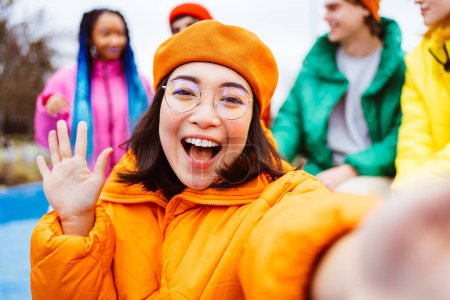 Foto de Grupo multirracial de jóvenes amigos felices que se reúnen al aire libre en invierno, vistiendo chaquetas de invierno y divirtiéndose, mujer asiática tomando selfie pov - Millennials multiétnicos que se unen en un área urbana, conceptos sobre la juventud y las liberaciones sociales - Imagen libre de derechos
