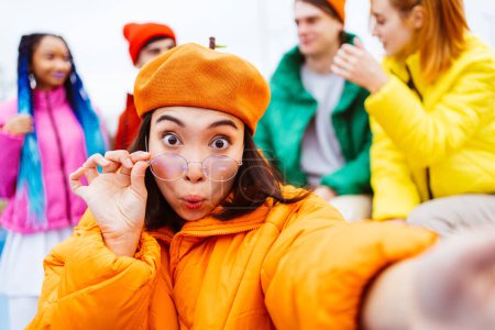 Photo for Multiracial group of young happy friends meeting outdoors in winter, wearing winter jackets and having fun, asian woman taking pov selfie - Multiethnic millennials bonding in a urban area, concepts about youth and social releationships - Royalty Free Image