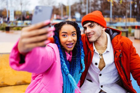 Téléchargez les photos : Multiracial jeune couple d'amoureux sortir ensemble en plein air en hiver en utilisant application de médias sociaux sur smartphone, porter des vestes d'hiver et avoir du plaisir - Multiethnique millennials collage dans une zone urbaine, concepts sur les jeunes et les relations sociales - en image libre de droit
