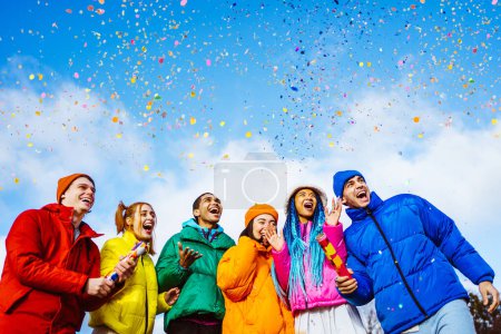 Téléchargez les photos : Groupe multiracial de jeunes amis heureux se réunissant à l'extérieur en hiver et célébrant la fête avec un tireur de confettis, portant des vestes d'hiver et s'amusant - Les milléniaux multiethniques se lient dans une zone urbaine, concepts sur les jeunes et les libérations sociales - en image libre de droit