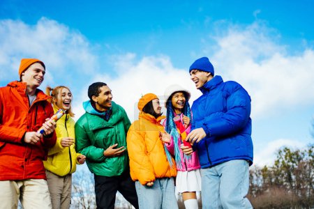 Foto de Grupo multirracial de jóvenes amigos felices que se reúnen al aire libre en invierno y celebran una fiesta con un tirador de confeti, vistiendo chaquetas de invierno y divirtiéndose. - Imagen libre de derechos