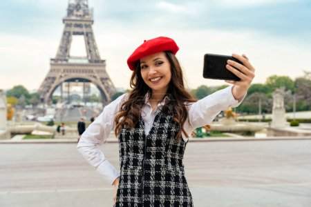 Foto de Beautiful young woman visiting paris and the eiffel tower. Parisian girl with red hat and fashionable clothes having fun in the city center and landmarks area - Imagen libre de derechos
