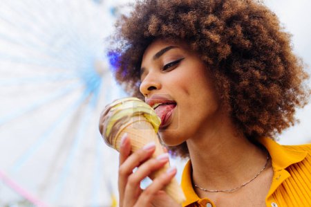 Photo for Beautiful young black woman eating ice cream at amusement park - Cheerful african-american female portrait during summertime vacation- Leisure, people and lifestyle concepts - Royalty Free Image