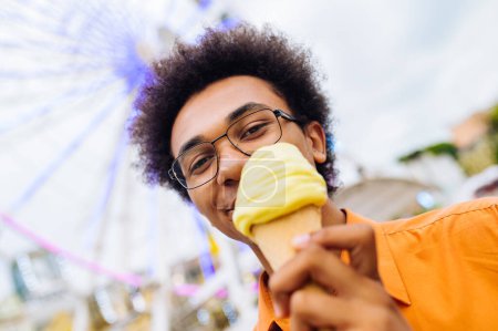 Téléchargez les photos : Handsome young black man eating ice cream at amusement park - Cheerful african-american male portrait during summertime vacation- Leisure, people and lifestyle concepts - en image libre de droit