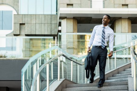 Téléchargez les photos : Charmant homme d'affaires afro-américain PDG dans un élégant costume élégant d'entreprise dans un centre d'affaires en plein air noir banlieusard aller au travail, ville et quartier financier en arrière-plan - en image libre de droit
