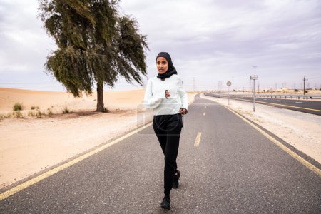 Téléchargez les photos : Belle femme arabe du Moyen-Orient portant un entraînement hijab à l'extérieur dans une zone désertique - Sportive sportive musulmane adulte portant des vêtements de sport en burkini faisant de l'exercice de fitness - en image libre de droit