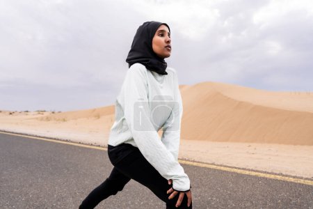Foto de Hermosa mujer árabe del medio oriente que usa entrenamiento de hijab al aire libre en una zona desértica. Deportiva atlética musulmana adulta que usa ropa deportiva de burkini que hace ejercicio físico. - Imagen libre de derechos