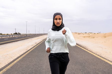 Téléchargez les photos : Belle femme arabe du Moyen-Orient portant un entraînement hijab à l'extérieur dans une zone désertique - Sportive sportive musulmane adulte portant des vêtements de sport en burkini faisant de l'exercice de fitness - en image libre de droit