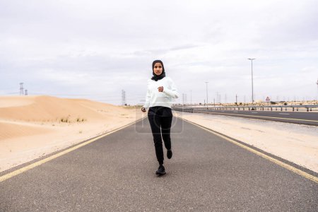 Foto de Hermosa mujer árabe del medio oriente que usa entrenamiento de hijab al aire libre en una zona desértica. Deportiva atlética musulmana adulta que usa ropa deportiva de burkini que hace ejercicio físico. - Imagen libre de derechos