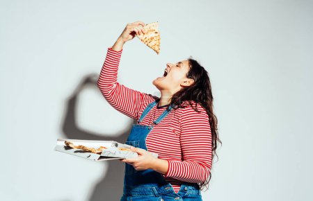 Téléchargez les photos : Modèle femme taille plus posant en studio et s'amusant. Manger une pizza italienne de la boîte - en image libre de droit
