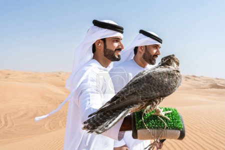 Foto de Dos hombres de Oriente Medio que llevan puesto el tradicional emirati árabe kandura vinculación en el desierto y la celebración de un pájaro halcón - amigos musulmanes árabes se reúnen en las dunas de arena en Dubai - Imagen libre de derechos