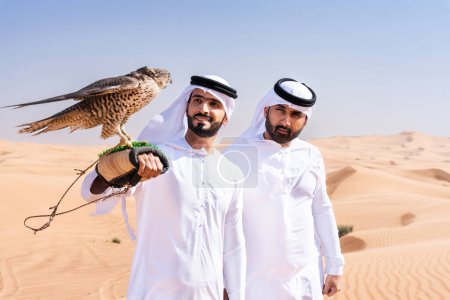 Téléchargez les photos : Deux hommes du Moyen-Orient portant des kandura arabes emirati traditionnels se lient dans le désert et tenant un oiseau de faucon - des amis musulmans arabes se rencontrent dans les dunes de sable de Dubaï - en image libre de droit