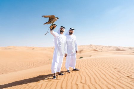 Foto de Dos hombres de Oriente Medio que llevan puesto el tradicional emirati árabe kandura vinculación en el desierto y la celebración de un pájaro halcón - amigos musulmanes árabes se reúnen en las dunas de arena en Dubai - Imagen libre de derechos