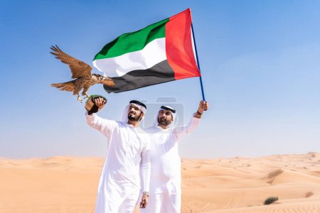 Foto de Dos hombres de Oriente Medio que llevan puesto el tradicional emirati árabe kandura vinculación en el desierto y la celebración de un pájaro halcón y la bandera de los Emiratos Árabes Unidos amigos musulmanes se reúnen en las dunas de arena en Dubai - Imagen libre de derechos