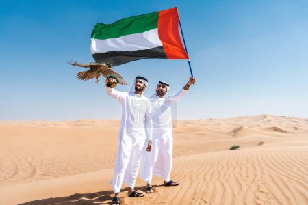Photo for Two middle-eastern men wearing traditional emirati arab kandura bonding in the desert and holding a falcon bird and UAE flag - Arabian muslim friends meeting at the sand dunes in Dubai - Royalty Free Image