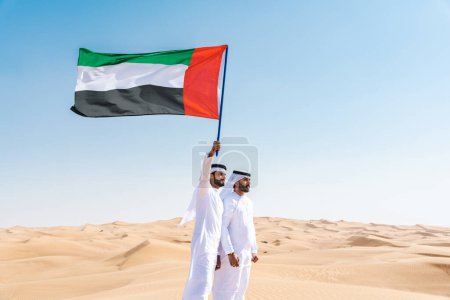 Foto de Dos hombres de Oriente Medio que llevan puesto el tradicional emirati árabe kandura vinculación en el desierto y la celebración de la bandera emirati - Amigos musulmanes árabes se reúnen en las dunas de arena en Dubai - Imagen libre de derechos