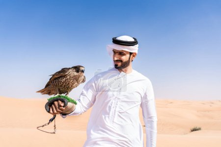 Photo for Middle-eastern man wearing traditional emirati arab kandura in the desert and holding a falcon bird - Arabian muslim adult person at the sand dunes in Dubai - Royalty Free Image
