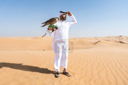 Photo for Middle-eastern man wearing traditional emirati arab kandura in the desert and holding a falcon bird - Arabian muslim adult person at the sand dunes in Dubai - Royalty Free Image